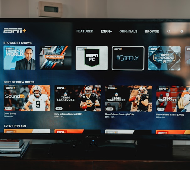 A television screen displaying the ESPN+ streaming interface with various sports-related shows and categories. The screen includes sections such as 'Browse by Shows' and 'Best of Drew Brees' with thumbnails of sports programs featuring athletes in action and analysts.