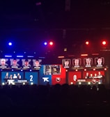 An esports event with a large screen displaying two teams, Team Kaliber and Rise Nation, competing against each other. The screen shows profiles of team members and scores, with Team Kaliber leading 2-0. The venue is dimly lit with blue and red lighting, and an audience can be seen watching.