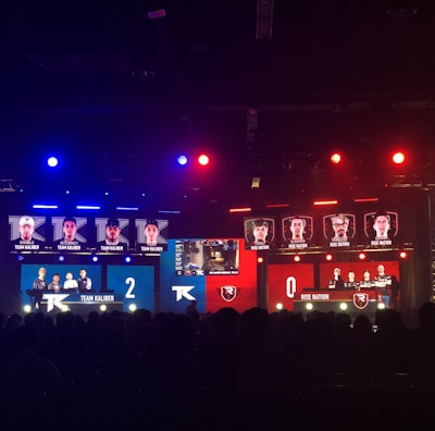 An esports event with a large screen displaying two teams, Team Kaliber and Rise Nation, competing against each other. The screen shows profiles of team members and scores, with Team Kaliber leading 2-0. The venue is dimly lit with blue and red lighting, and an audience can be seen watching.