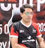 A man with glasses is sitting in a gaming chair, focused on a monitor in front of him. He is wearing a black and red esports jersey with various logos. The background features a large banner with text and numbers in bold fonts.