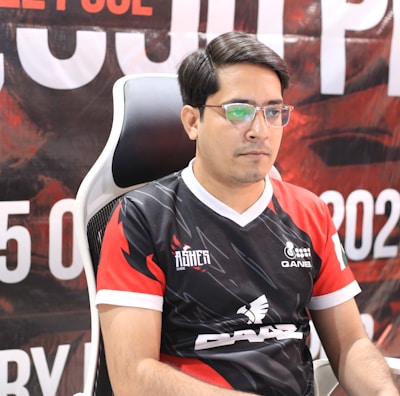 A man with glasses is sitting in a gaming chair, focused on a monitor in front of him. He is wearing a black and red esports jersey with various logos. The background features a large banner with text and numbers in bold fonts.